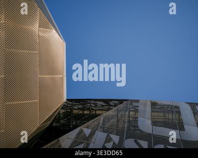 Abstract architectural detail of a modern building exterior featuring geometric golden perforated metal cladding intersecting with glass panels agains Stock Photo