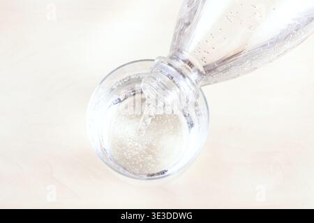 View from above of pouring mineral water from a clear plastic bottle into glass on a light brown table from close range Stock Photo