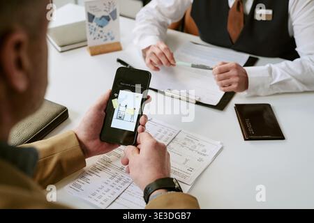 Middle Aged Caucasian Man Using Smartphone While Consulting Bank Employee Stock Photo