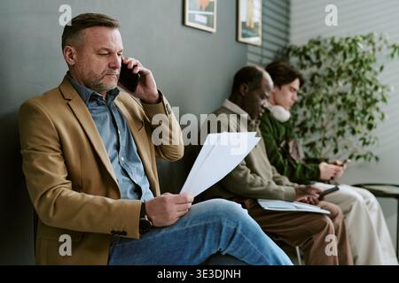 Middle Aged Caucasian Man Talking on Smartphone While Reading Documents in Bank Stock Photo