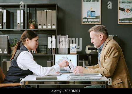 Young Adult Asian Woman Assisting Middle Aged Caucasian Man with Banking Documents Stock Photo