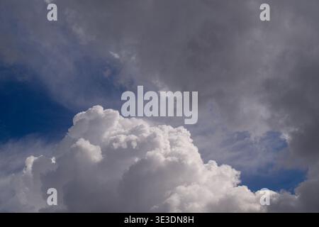 Large, fluffy white clouds dominate a deep blue sky. Dark gray storm clouds loom above, adding drama. Scene suggests impending weather change or calm Stock Photo