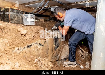 Man builder repairman drills hole in concrete block with perforator in basement of wooden house Stock Photo