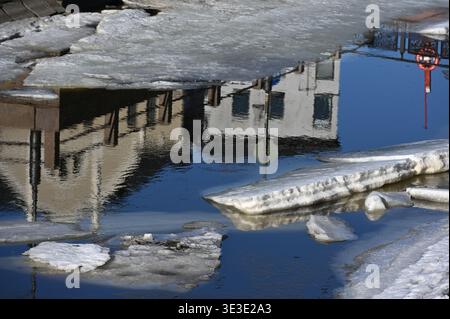Urban winter scene with melting ice floating on water, reflecting nearby buildings and street elements, capturing the transition between cold season a Stock Photo