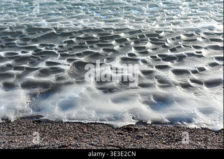 Frozen shoreline with textured melting ice and snow formations near a pebbled beach, illuminated by bright winter sunlight creating patterns and shado Stock Photo