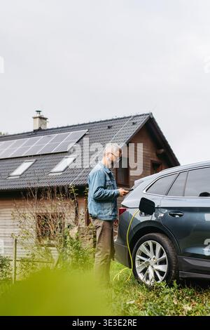 Charging electric car at home, using app. Solar panels on roof behind him. Stock Photo