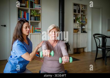 Physiotherapist assisting senior woman with dumbbell exercise, home rehabilitation. Stock Photo