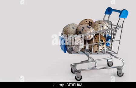 Mini shopping cart filled with speckled quail eggs on white background, symbolizing Easter shopping Stock Photo