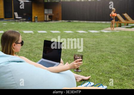Freelancer woman works on laptop in backyard garden with wine glass while son plays in background. Concept of remote work and relaxing summer Stock Photo