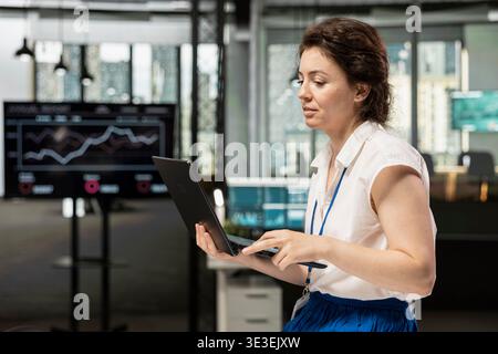 Female team leader stands in office and analyzes business analytics on laptop. Businesswoman managing company data, preparing reports to boost productivity and success in financial firm. Stock Photo