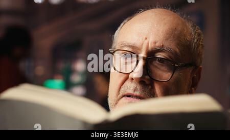 Old person with spectacles enjoys reading a book at the library, being happy and enjoying recreation. Aged man with book reading hobby, retirement leisure activity in quiet space. Stock Photo
