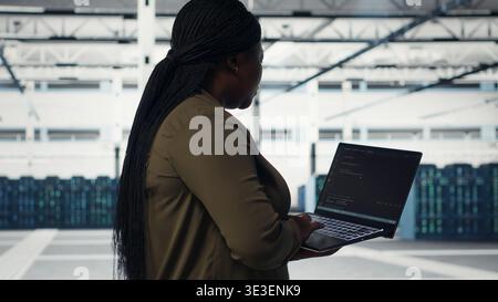 Technician with laptop walks through data center server rows, implementing advanced power management solutions. Worker using notebook to oversee server hub, reducing energy consumption, camera B Stock Photo