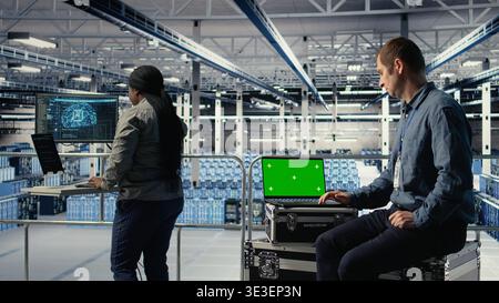 Data center IT experts investigate AI configurations using green screen laptop, making adjustments. Server hub engineers check deep learning errors on chroma key notebook, camera A Stock Photo