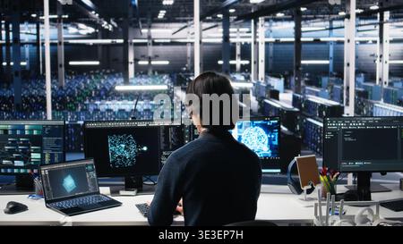 Data center system administrator managing AI systems for uptime and efficiency. IT employee using artificial intelligence technology on computer and laptop in server hub, camera A Stock Photo