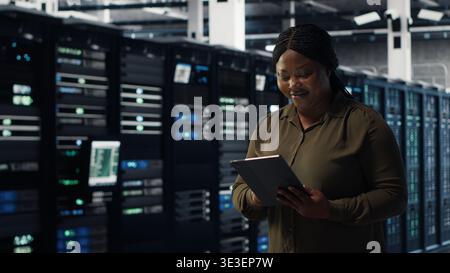 Engineer with tablet walks through data center server rows, implementing advanced power management solutions. IT professional using device to oversee server hub, reducing energy consumption, camera B Stock Photo