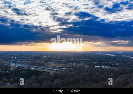 Aerial view near Warsaw at sunset shows white townhouses, farms, leafless forests, fields, and a winding river on the right under layered blue storm c Stock Photo