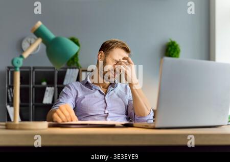 Tired accountant rubbing eyes by laptop while handling deadlines and complex audit Stock Photo