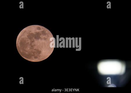 A full moon shines brightly in the dark night sky with a blurred bright light source in the foreground creating contrast between natural and artificia Stock Photo