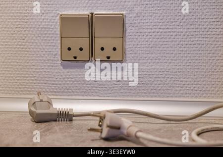 Beige electrical outlets mounted on a textured wall with two unplugged power cords lying on the floor beneath them, showing a typical indoor electrica Stock Photo