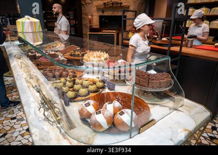 Milan, Italy - 9 Mar 24: Assorted cakes and pastries displayed in a glass case at the Starbucks Reserve Roastery in Milan, Italy, with staff working b Stock Photo