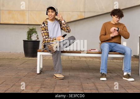 Diverse male students sitting on wooden bench in courtyard, wearing headphones and holding phone Stock Photo