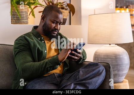 African American adult sitting on gray sofa in lounge wearing mustard sweater holding blue phone Stock Photo