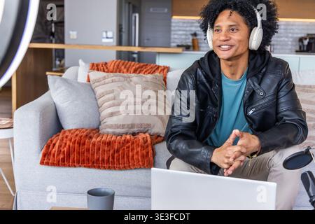 African American man late-20s sitting on gray sofa in leather jacket, recording with laptop and mic Stock Photo