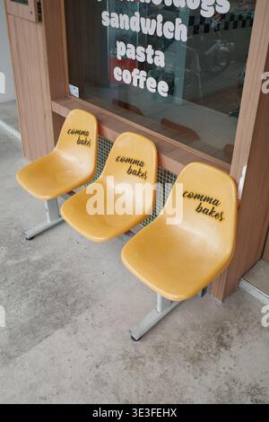 Yellow plastic chairs sit outside a cafe. They have "bakes" written on them. The window shows food offerings. Simple, clean, and inviting seating. Ide Stock Photo