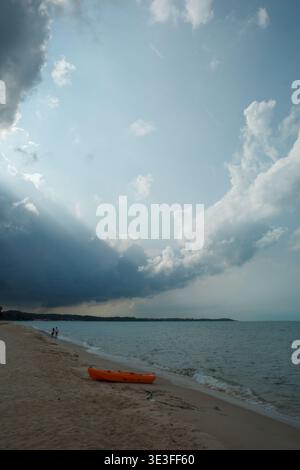Orange kayak rests on sandy shore. Dark clouds loom over calm ocean waters. Lone figure walks near water's edge. Sunlight pierces cloud layer above. S Stock Photo