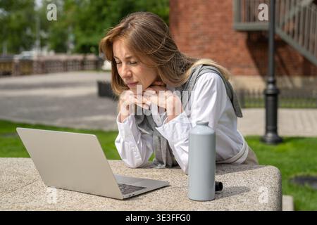 Curious positive businesswoman works on laptop outdoors, focused lady watching webinar on computer. Stock Photo