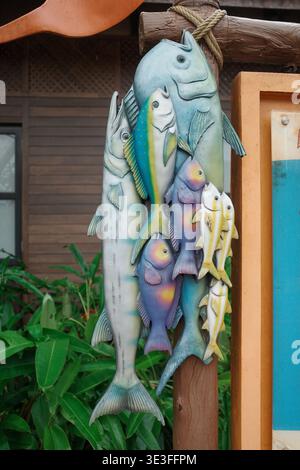 Colorful fish sculptures hang from a wooden post. Green foliage frames the background. Wooden structure and blue panel beside the display. Fish vary i Stock Photo