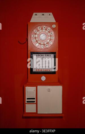 Vintage wall-mounted payphone in red and white. Rotary dial and coin slot visible. Clear glass panel displays instructions. Simple design with retro a Stock Photo