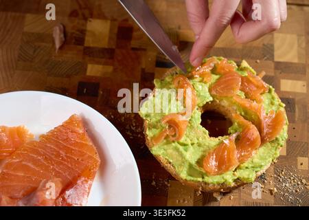 Hand places smoked salmon on avocado bagel half on wooden board. Concept of healthy breakfast preparation and homemade brunch recipe. Stock Photo
