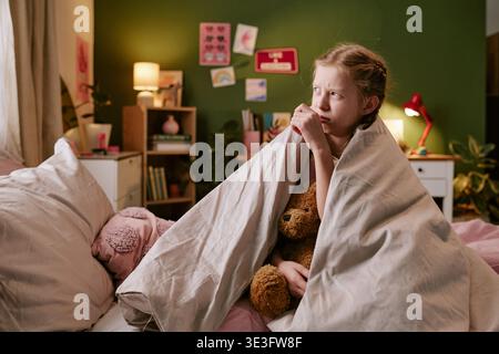 Girl Hugging Toy Bear While Sitting in Bed Looking Worried Stock Photo