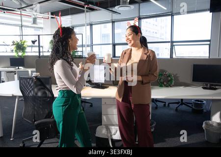 Diverse female coworkers chatting and holding paper cups, wearing holiday headbands at office Stock Photo