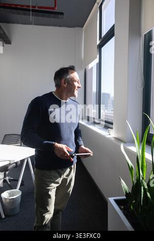 Middle-aged man standing by office windowsill holding tablet, wearing navy sweater, khaki trousers Stock Photo