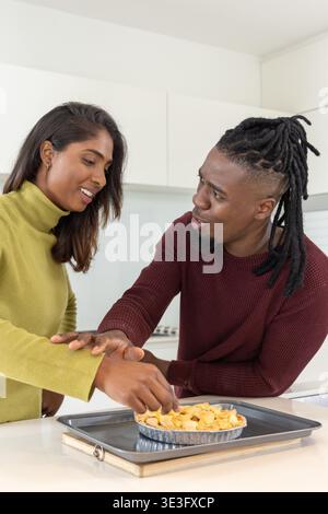 Diverse couple in green turtleneck and burgundy knit preparing pie on kitchen island with foil pan Stock Photo