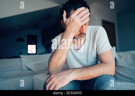 Tired man resting head in hand while sitting on couch in dim living room, concept of exhaustion, worry or burnout, selective focus Stock Photo