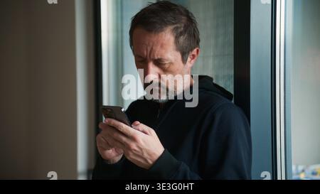 Mature bearded man leaning on apartment window while typing message on smartphone, casual everyday communication at home, selective focus Stock Photo