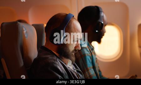 Passenger in airplane seat using laptop to work on flight, feeling tired about working on vacation. Flying on plane during sunset hour, person travelling with international airline. Stock Photo