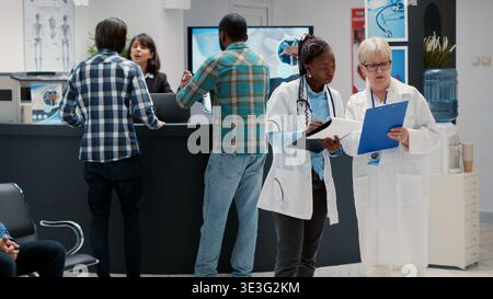 Diverse medical team analyzing checkup report files in waiting area lobby, talking about consultation. Doctors discussing about healthcare service, looking at documents in hospital reception. Stock Photo