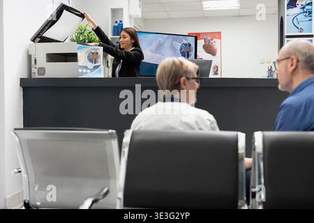 Asian receptionist using copier and scanning machine at front desk of modern healthcare facility. Female hospital secretary handles printing of medical forms as elderly doctor consults with patient. Stock Photo