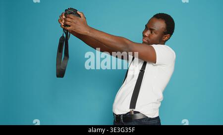 African american person holding dslr camera to take pictures, standing in studio over blue background. Photographer using professional equipment to capture photos, zoom focus snap. Stock Photo