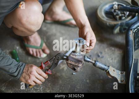 Focused mechanic fixing metal part of small bicycle using pliers dirty floor working intently repair shop fixing mechanical equipment carefully concen Stock Photo