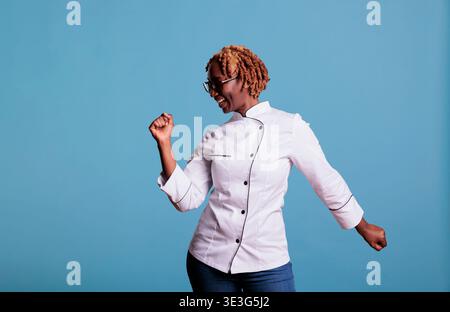 Cheerful african american baker in uniform and dancing with excitement in front of camera. Energetic female cook unleashing moves to the rhythm of the music in studio shot. Stock Photo