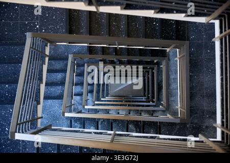 An overhead perspective of a contemporary architectural spiral stairwell, showcasing dark terrazzo steps and geometric metal railings. The repeated re Stock Photo