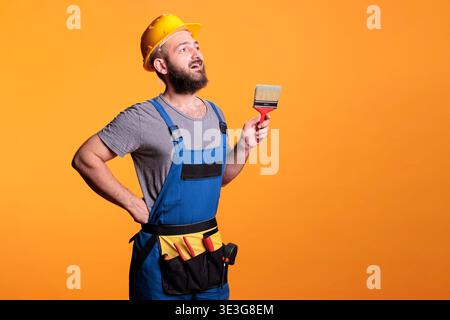 Young painter decorator painting walls with brush in studio, posing with paintbrush and tools before working on renovation. Craftsman expert using brush and color to do diy project. Stock Photo