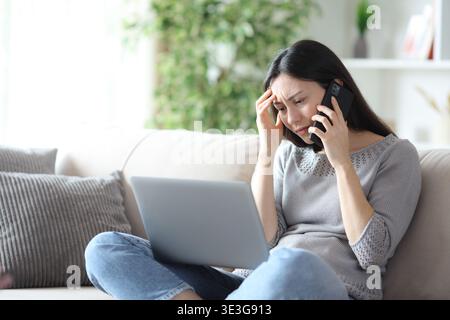 Worried asian woman checking laptop and talking on phone sitting on a couch at home Stock Photo