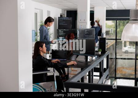 Asian disabled software programmer writing html code in agency big data room, using security system. Engineer with physical disability programming user interface artificial intelligence office. Stock Photo