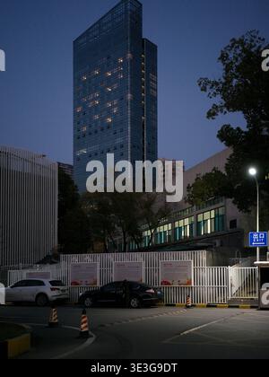 Wuhan, China - March 21, 2025: A modern glass skyscraper rising behind a white metal fence and a parking lot. The image shows parked cars and urban ar Stock Photo